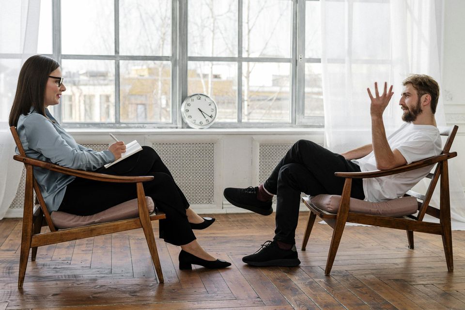 Two people are sitting in chairs facing each other, having a conversation in a bright room.