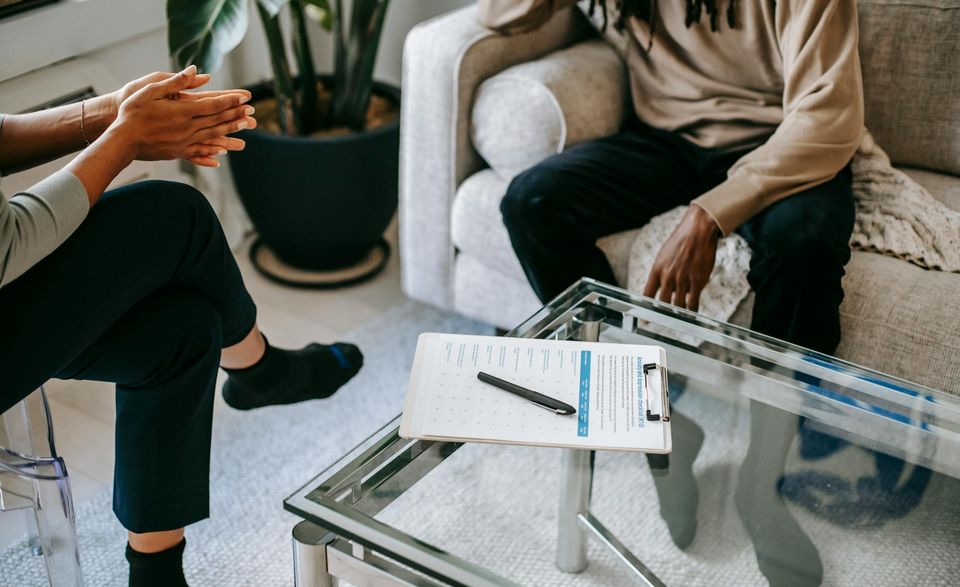 Two people are sitting and talking in a living room, with a clipboard and pen on a glass coffee table between them. Two people are sitting and talking in a living room, with a clipboard and pen on a glass coffee table between them.