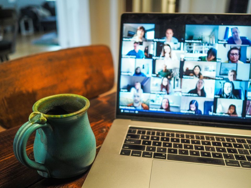A laptop displaying a video conference call is placed on a wooden table next to a green ceramic mug. A laptop displaying a video conference call is placed on a wooden table next to a green ceramic mug.